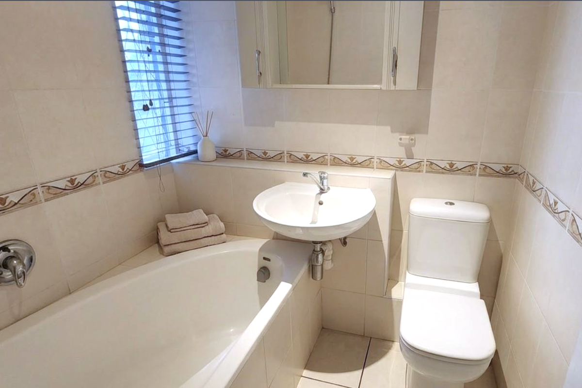 A functional bathroom featuring a bathtub, pedestal basin, toilet, and neutral tiled finishes with a medicine cabinet and window blinds.