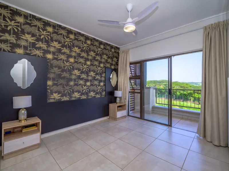 A well-maintained bedroom featuring tiled flooring, a ceiling fan, and sliding glass doors that open onto a private balcony with scenic views.