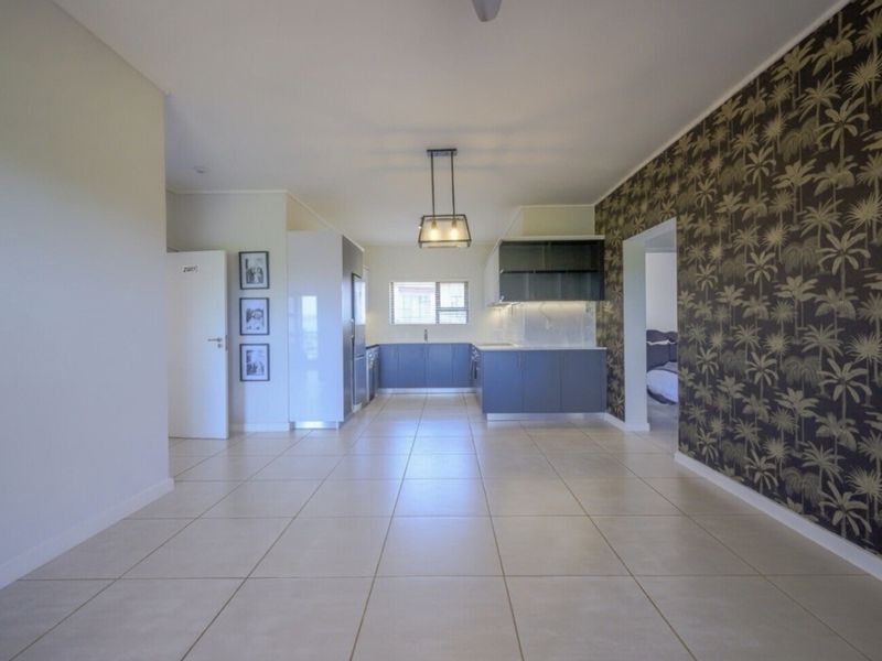 Spacious open-plan living and kitchen area featuring modern blue cabinetry, tiled flooring, and a decorative feature wall, offering a clean and versatile living space.