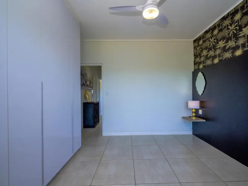 A spacious bedroom featuring neutral tiled flooring, extensive built-in cupboards, a ceiling fan, and a decorative feature wall with a floating shelf.
