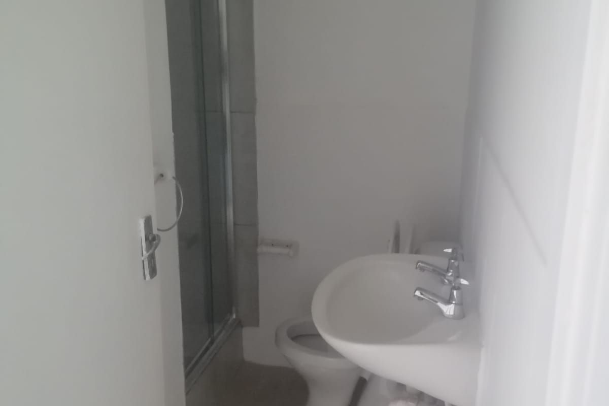A compact bathroom featuring a shower enclosure, pedestal basin, and toilet with neutral tiled flooring.