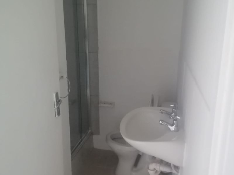 A compact bathroom featuring a shower enclosure, pedestal basin, and toilet with neutral tiled flooring.