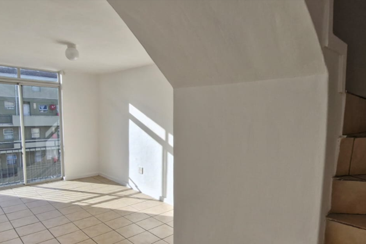 An empty living area featuring tiled flooring, a sliding glass door leading to a balcony, and an adjacent staircase with ceramic tiling.