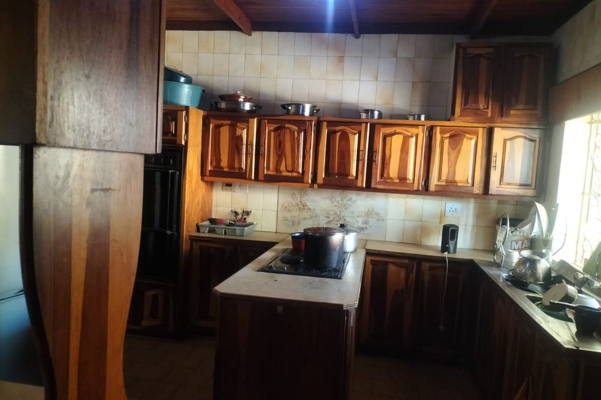 A dated kitchen featuring extensive wooden cabinetry, exposed timber ceiling beams, a built-in oven, and a central island with a hob.