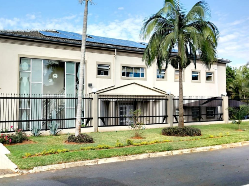 Front exterior view of a multi-story apartment building featuring solar panels, large glass windows, and secure perimeter fencing.