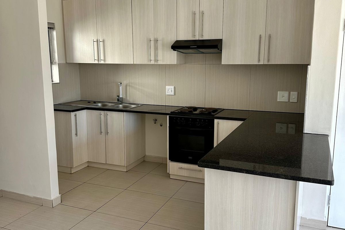 A clean, modern kitchen featuring light wood-grain cabinetry, dark granite countertops, built-in oven, hob, and extractor fan with neutral tiled flooring.