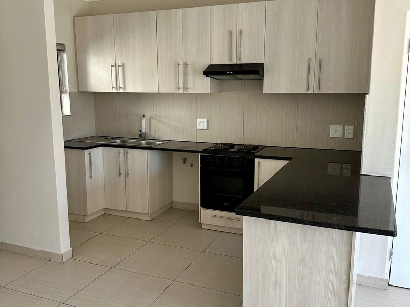 A clean, modern kitchen featuring light wood-grain cabinetry, dark granite countertops, built-in oven, hob, and extractor fan with neutral tiled flooring.