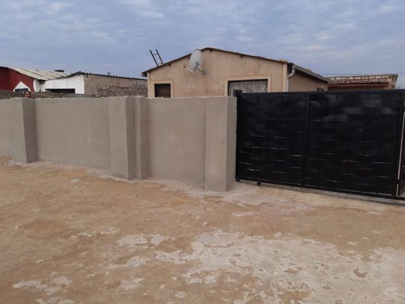 Front view of a residential property featuring a perimeter wall and a black sliding gate, with an unpaved driveway area.