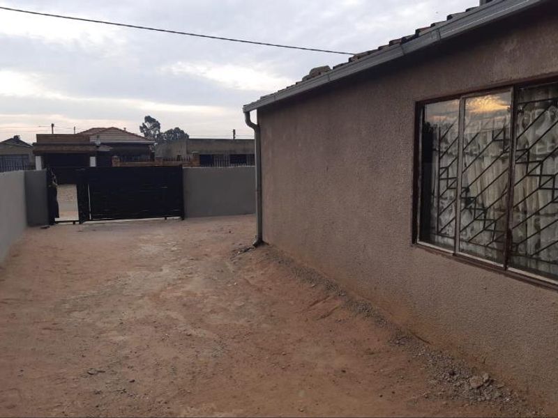 Rear exterior view of the property featuring a plain wall, sliding window with security bars, and an unpaved yard area with boundary wall and gate.