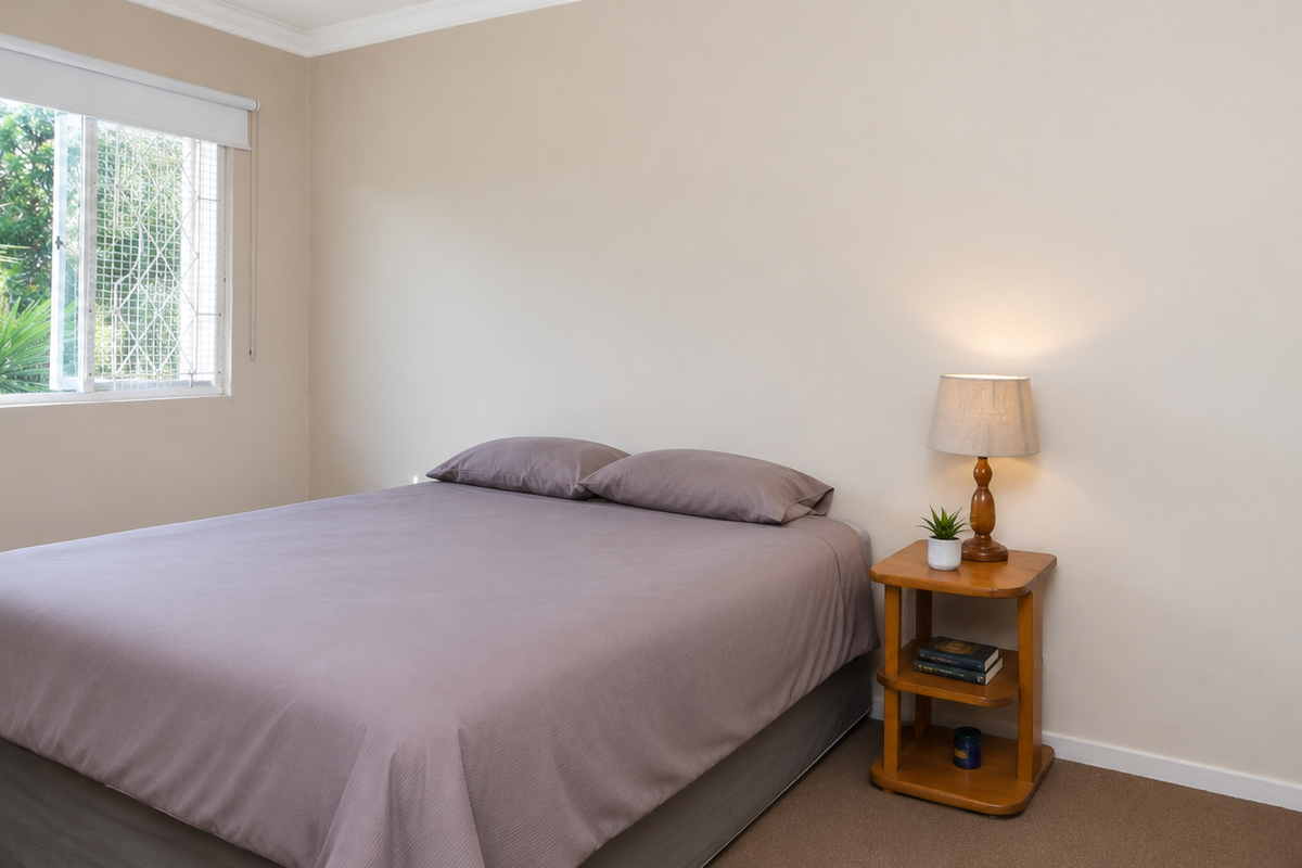 A bedroom featuring neutral walls, carpeted flooring, a built-in wardrobe, and a window fitted with security bars and a roller blind.