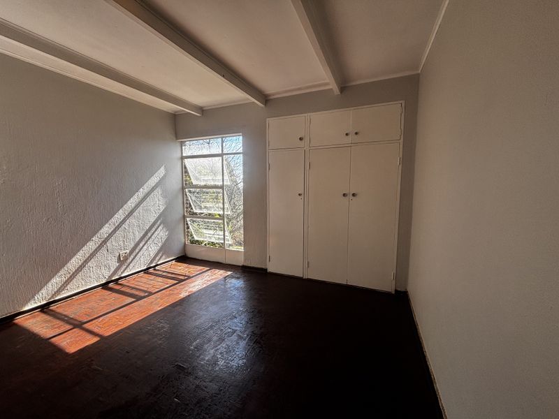 A bright, empty bedroom featuring original parquet flooring, built-in cupboards, and exposed ceiling beams, offering a blank canvas for renovation.