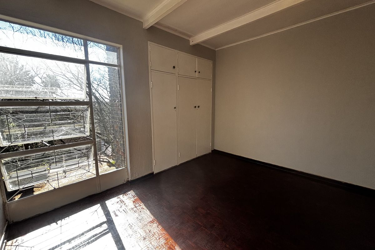 An empty bedroom featuring original parquet flooring, built-in cupboards, and a large window providing natural light.
