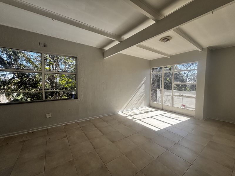 A bright, empty living area featuring tiled floors, exposed ceiling beams, and large windows that provide ample natural light and garden views.