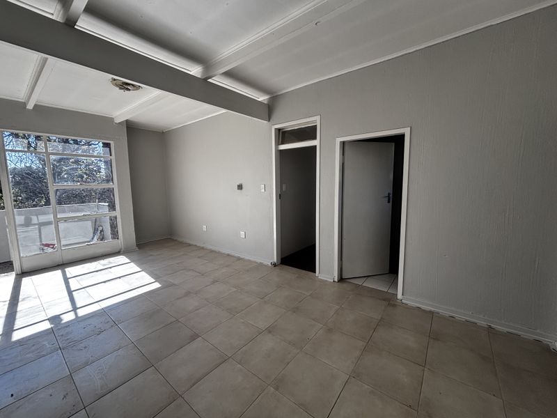An empty living room featuring tiled flooring, exposed beam ceilings, and a large window providing natural light and access to an outdoor area.