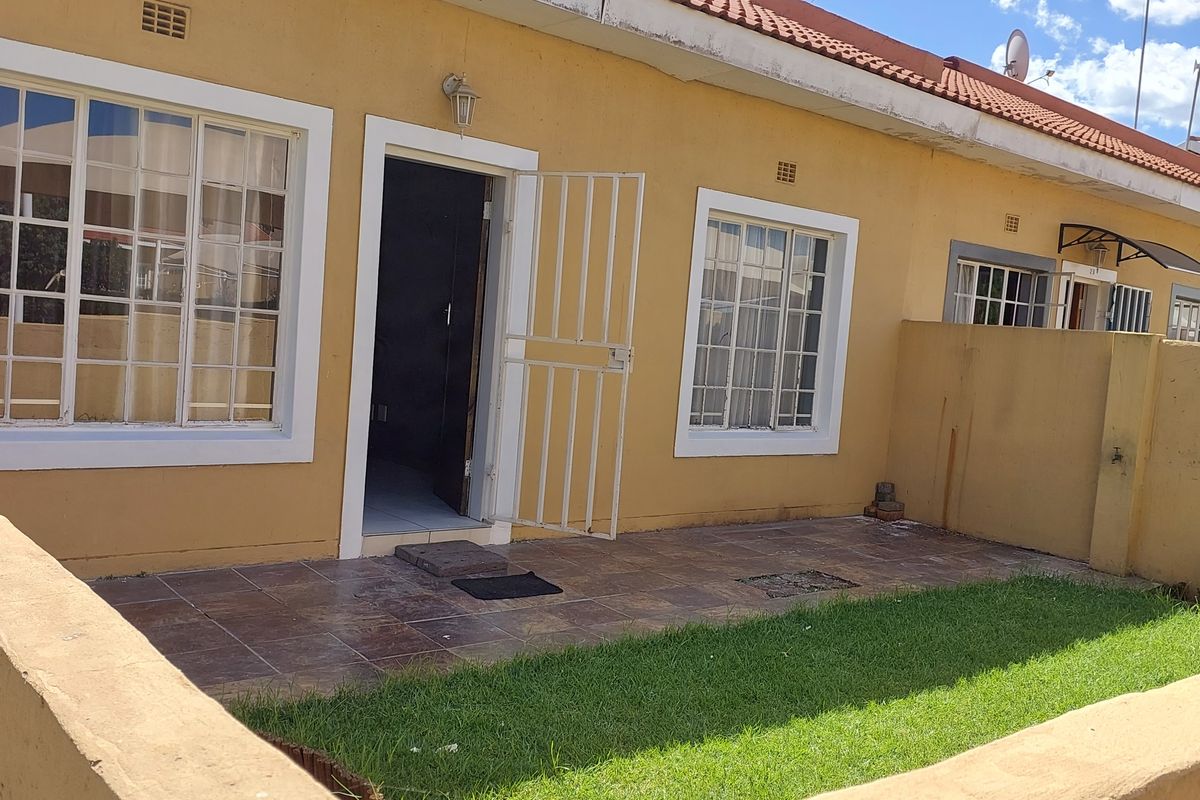 Rear exterior view of a townhouse featuring a tiled patio, small private lawn, security gate, and terracotta roof tiles.