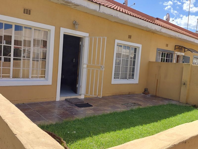 Rear exterior view of a townhouse featuring a tiled patio, small private lawn, security gate, and terracotta roof tiles.