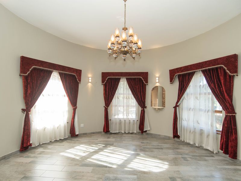 A spacious, empty living room featuring tiled flooring, a decorative chandelier, wall sconces, and large windows with heavy drapery.