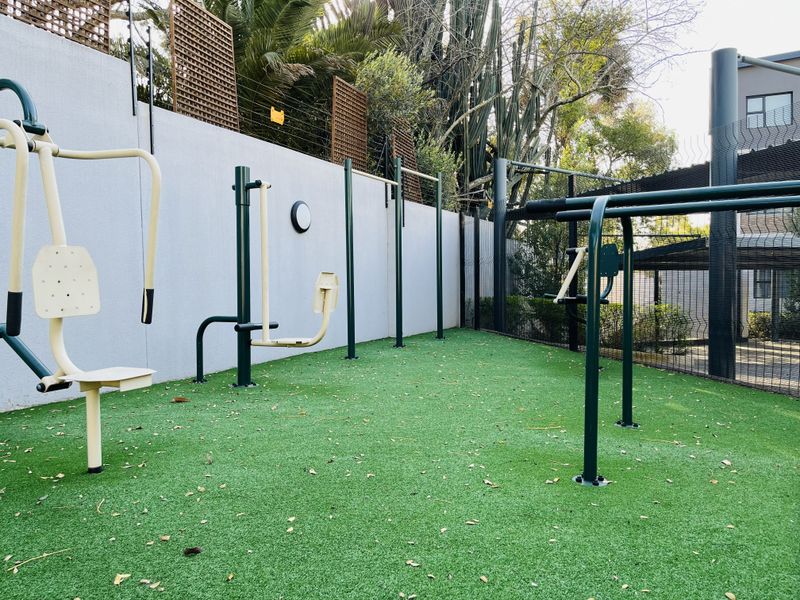 An outdoor gym area featuring fixed exercise equipment installed on artificial turf, enclosed by a perimeter wall and security fencing.
