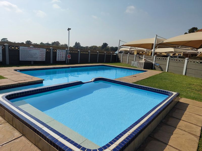 Communal swimming pool area featuring a main pool, a smaller wading pool, shade sails for sun protection, and paved surrounds.