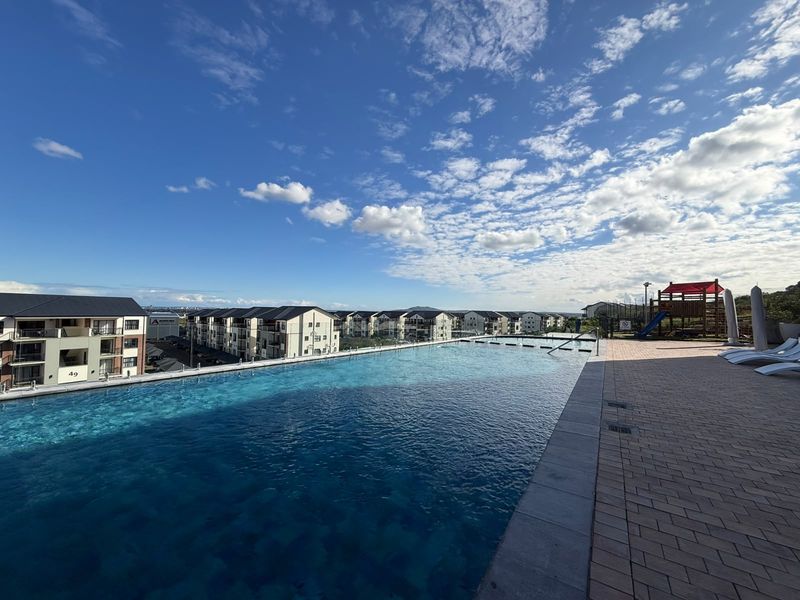 A well-maintained infinity edge swimming pool with a glass balustrade and paved deck, offering expansive views of the surrounding residential complex.