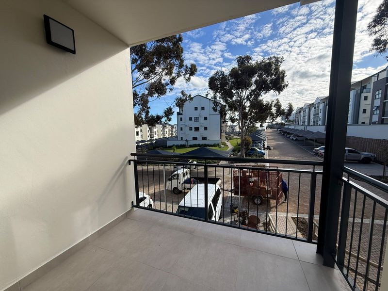 A clean, covered balcony featuring tiled flooring and a metal safety railing, overlooking the apartment complex grounds and parking area.