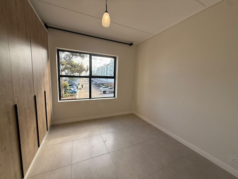 A bright, empty bedroom featuring modern tiled flooring, built-in wooden cupboards, and a large window with views of the complex parking area.