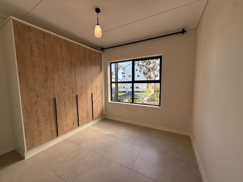 A bright, modern bedroom featuring large-format floor tiles, a spacious built-in wardrobe with wood-grain finish, and a black-framed window.