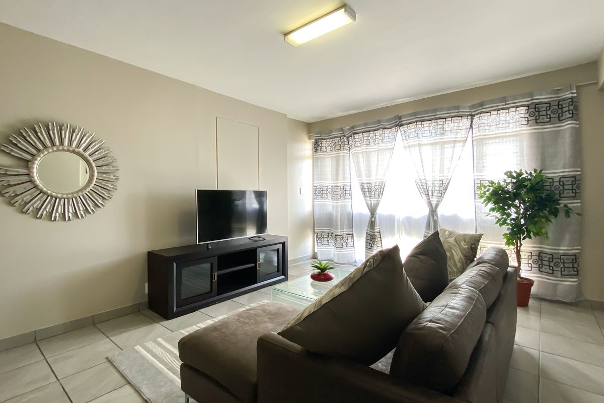 A comfortable living room featuring neutral wall tones, tiled flooring, and large windows with curtains allowing for natural light.