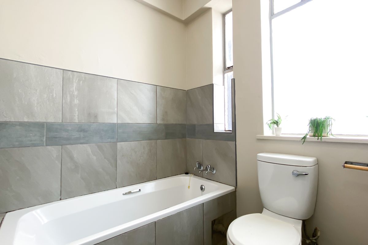 A functional bathroom featuring a bathtub with grey tiled surround, a standard toilet, and natural light from a window.