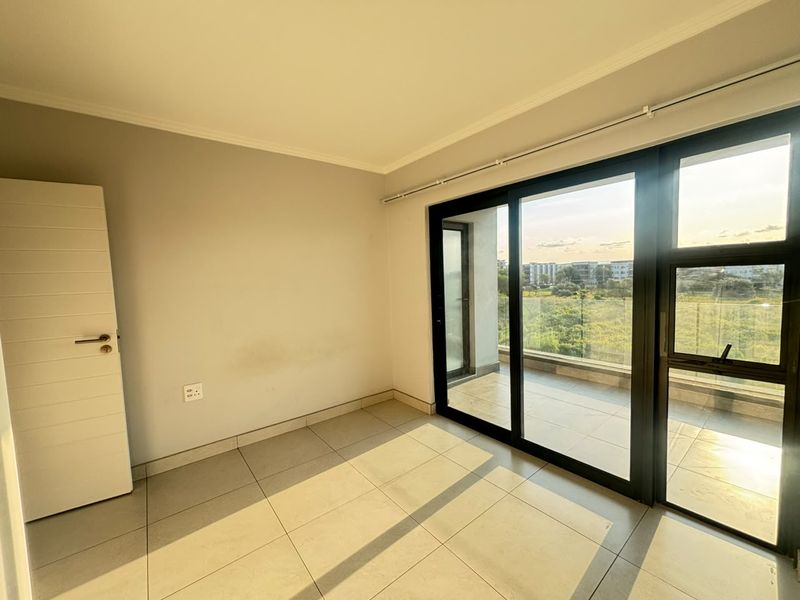 A bright, empty bedroom featuring large sliding glass doors that open onto a private balcony, offering natural light and tiled flooring throughout.