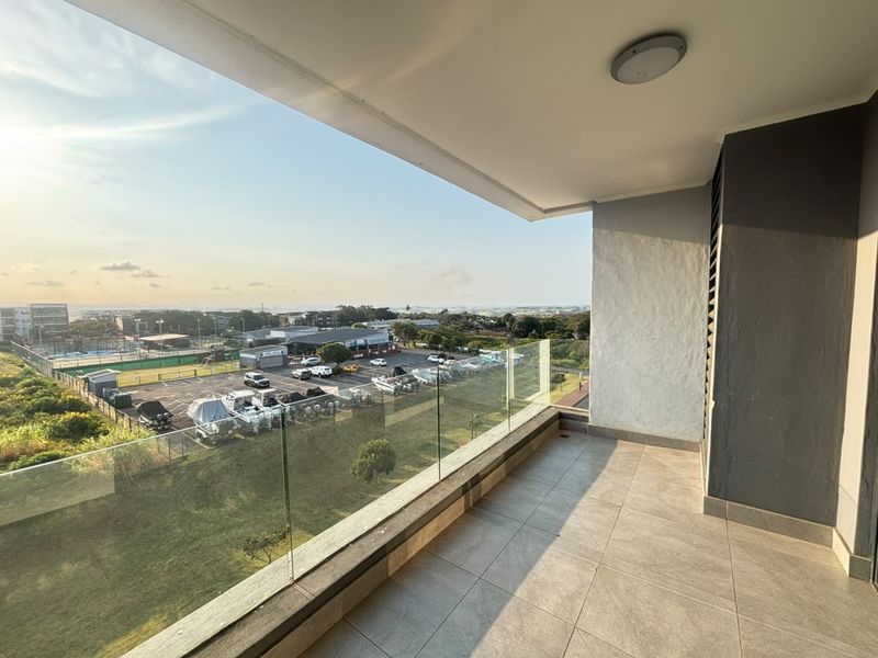 A spacious covered balcony featuring a modern glass balustrade and tiled flooring, offering an elevated view of the surrounding area.