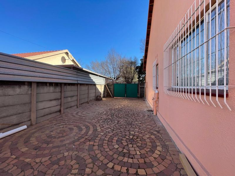 A paved rear exterior area featuring a precast concrete boundary wall, metal security gate, and window security bars on the side of the house.