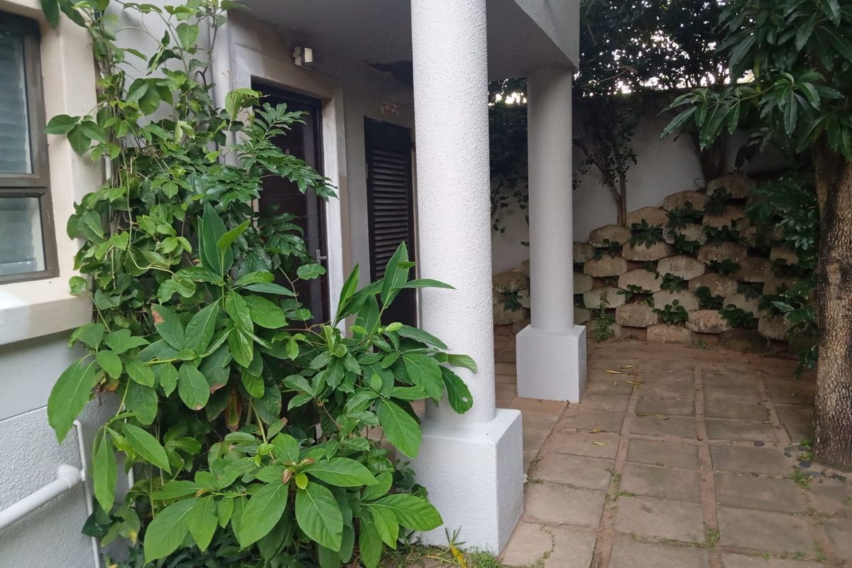 A rear patio area featuring paved flooring, structural columns, and a stone retaining wall, surrounded by established garden greenery.