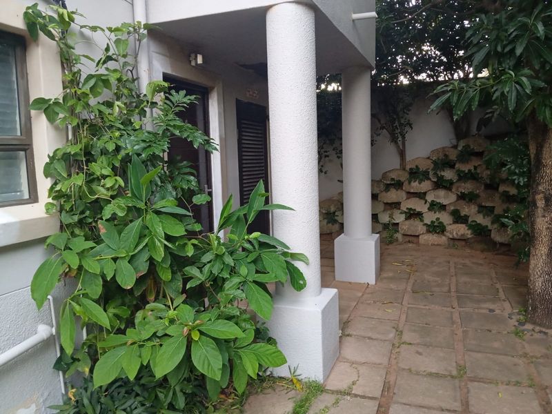 A rear patio area featuring paved flooring, structural columns, and a stone retaining wall, surrounded by established garden greenery.