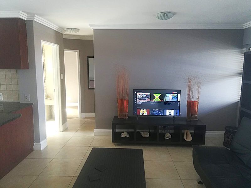 A living room area featuring tiled flooring, crown molding, and a view into the adjacent kitchen and hallway.