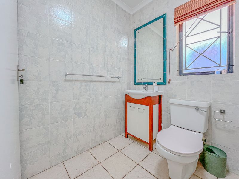A functional bathroom featuring a vanity cabinet with basin, wall-mounted mirror, toilet, and full-height wall tiling with security bars on the window.