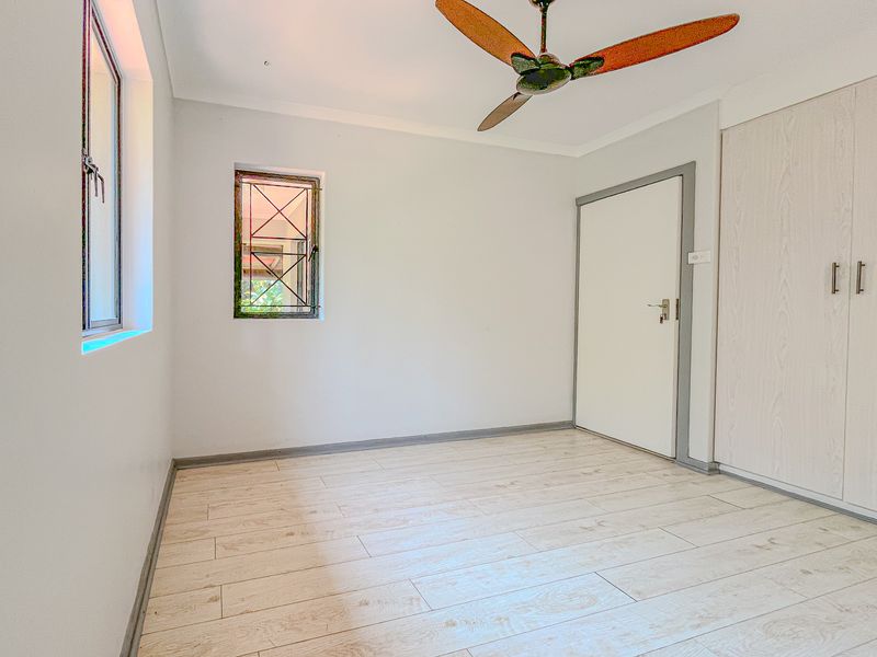 A bright, empty bedroom featuring light-toned tiled flooring, a ceiling fan, and built-in cupboards with security bars on the window.