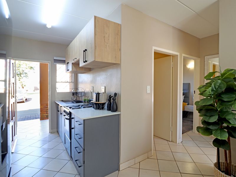 A compact, modern kitchen featuring grey lower cabinets, light wood upper cabinetry, stone countertops, and a built-in oven and hob, with tiled flooring throughout.