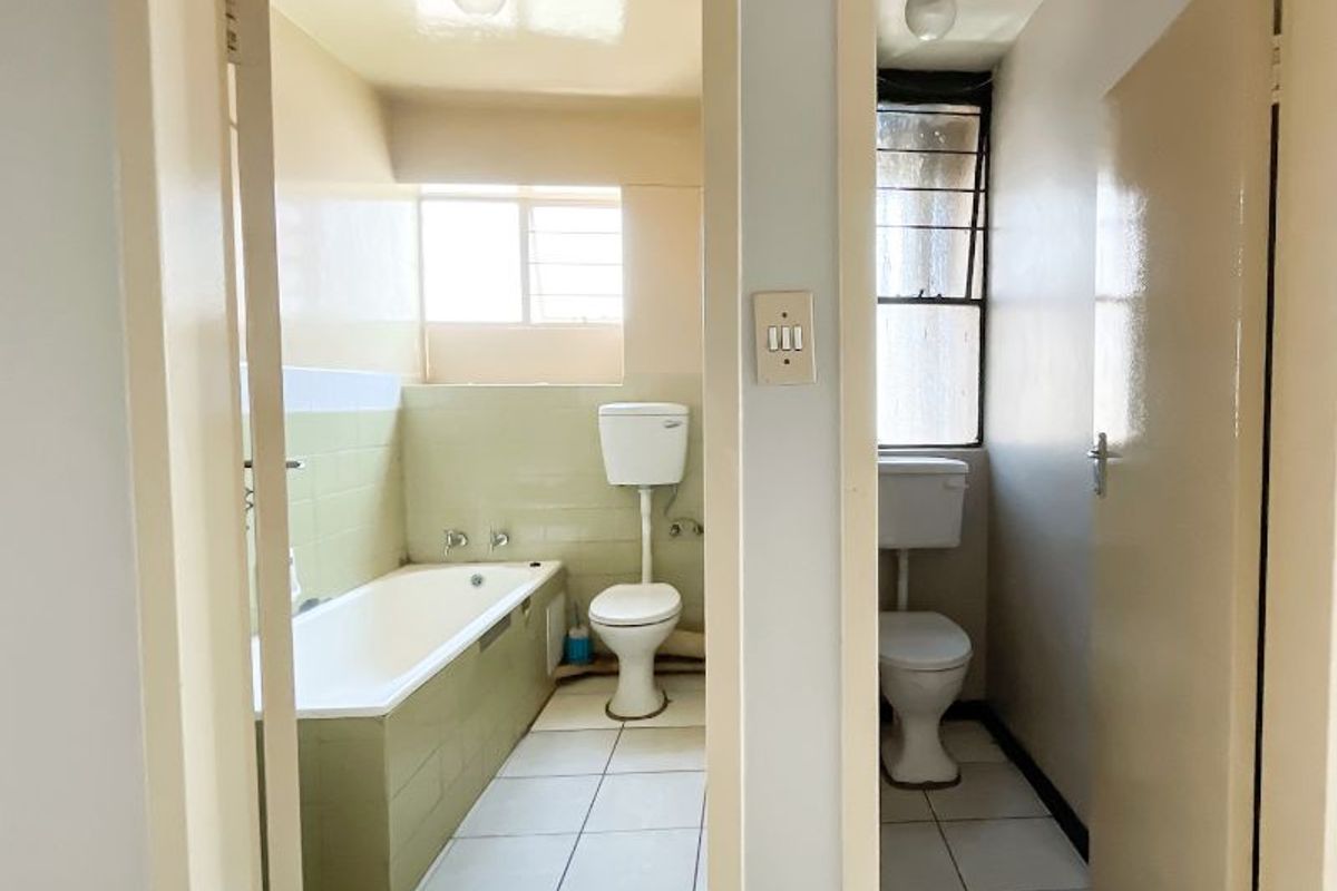A dated bathroom and separate toilet area featuring original tile finishes, standard sanitaryware, and natural light through windows with security bars.