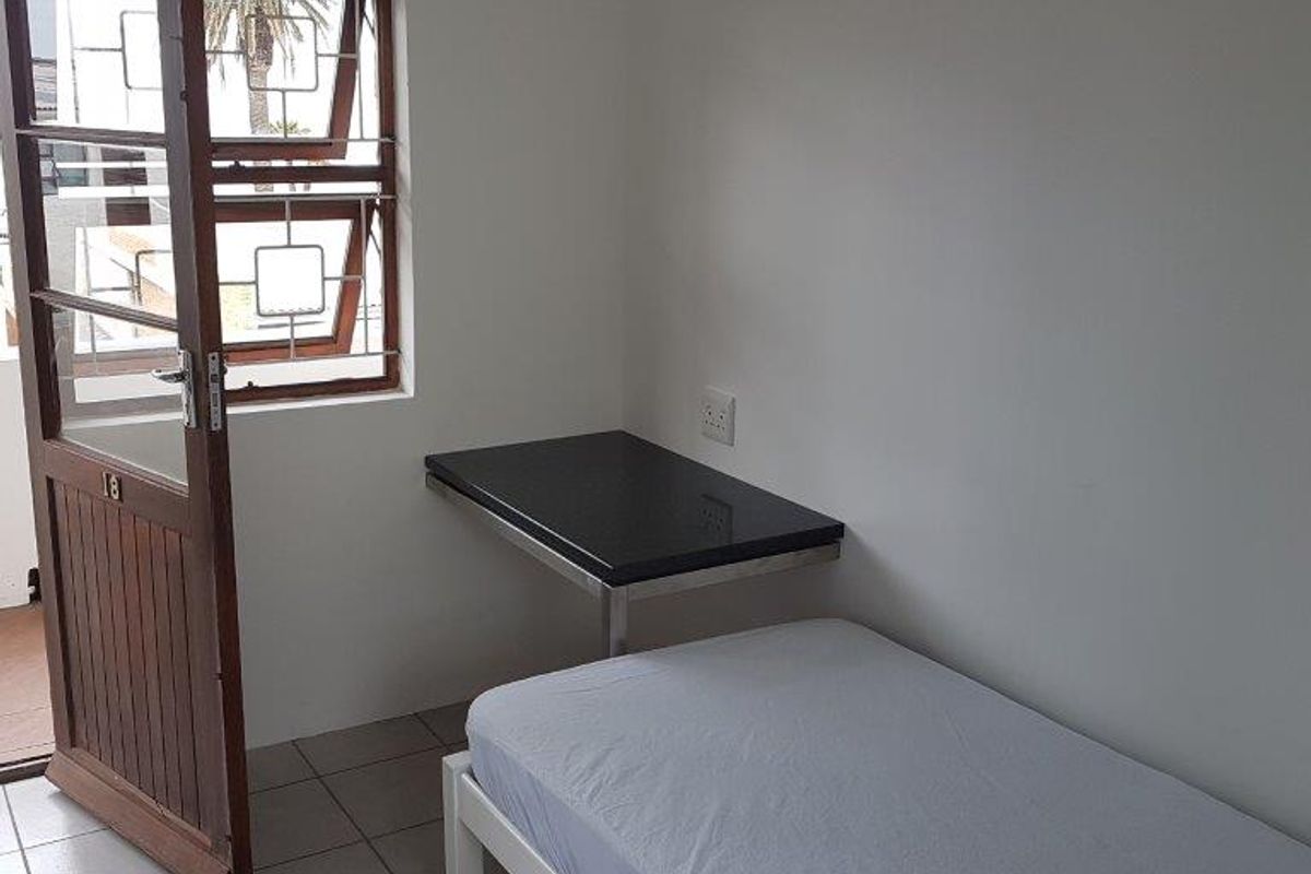 A compact bedroom featuring tiled flooring, a wall-mounted desk, and direct access to an outdoor area through a timber door with security bars.