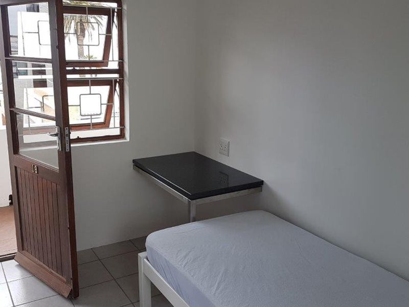 A compact bedroom featuring tiled flooring, a wall-mounted desk, and direct access to an outdoor area through a timber door with security bars.