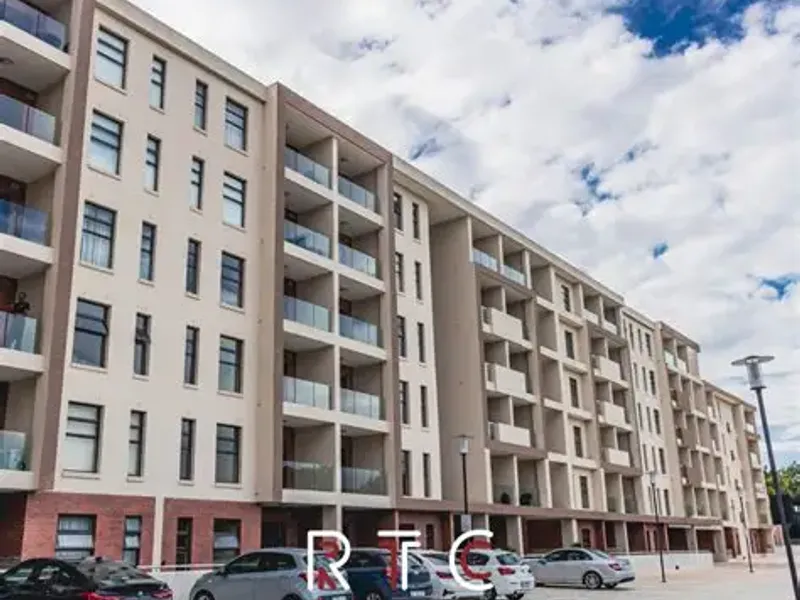 A modern multi-story apartment complex featuring contemporary architecture with private balconies, glass balustrades, and a paved ground-level parking area.