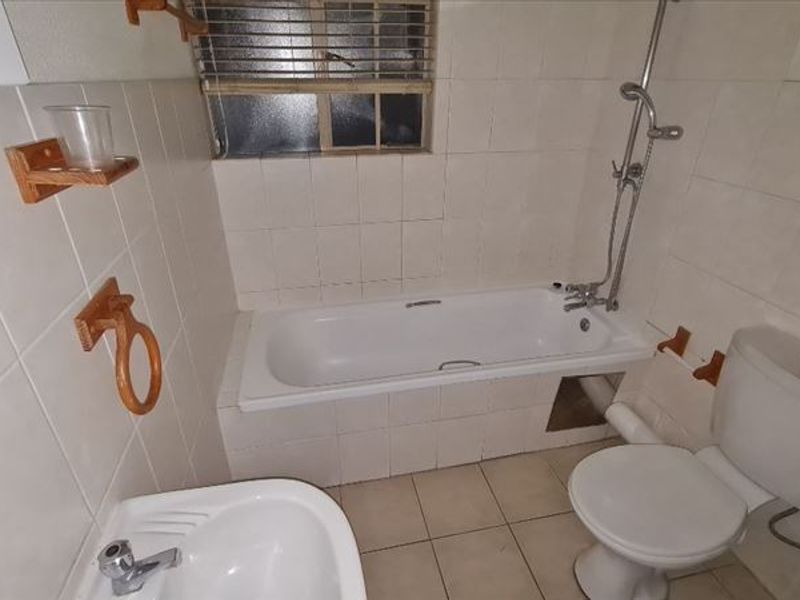 A functional bathroom featuring a bathtub with shower mixer, pedestal basin, and toilet, finished with neutral wall and floor tiling.