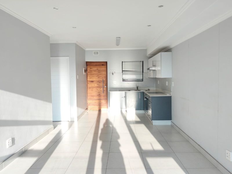 An open-plan living and kitchen area featuring tiled flooring, modern cabinetry, built-in appliances, and a timber entrance door, bathed in natural light.