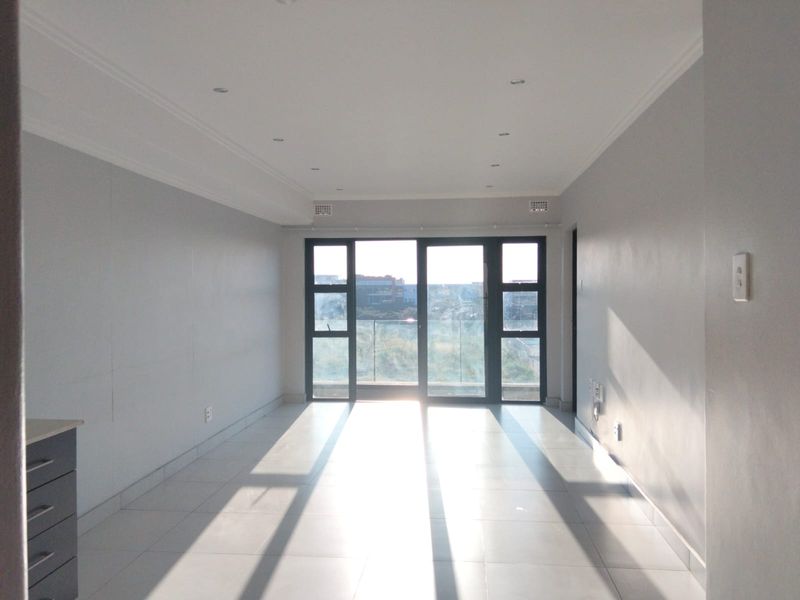 A bright, empty living area featuring neutral tiled flooring, recessed lighting, and large sliding glass doors leading to a balcony with a view.