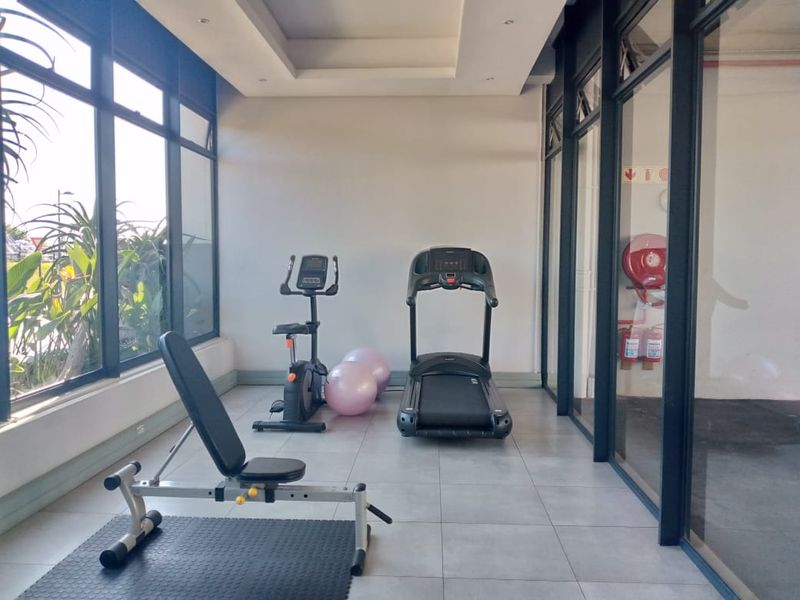 A bright, modern gym space featuring large glass windows, tiled flooring, and a recessed ceiling, equipped with essential fire safety infrastructure.