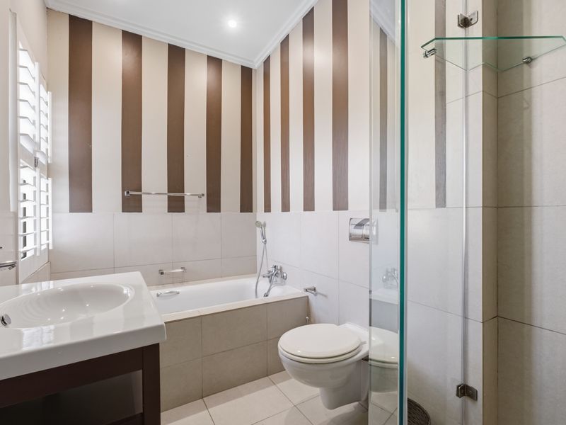 A modern bathroom featuring a built-in bathtub, wall-mounted toilet, glass shower enclosure, and vanity basin with neutral tiling and decorative striped wallpaper.
