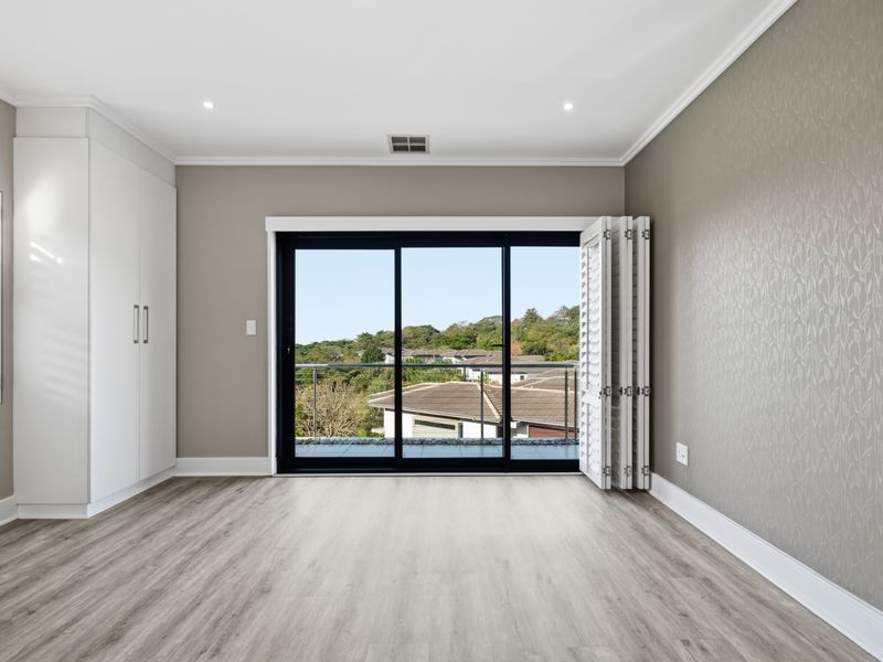 A bright, modern bedroom featuring built-in cupboards, laminate flooring, and large sliding doors opening to a balcony with scenic views.
