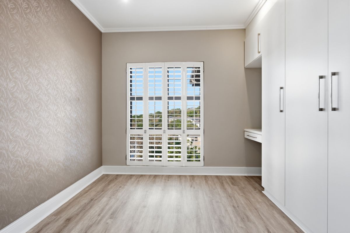 A bright, empty bedroom featuring modern laminate flooring, built-in cupboards with an integrated desk, and white plantation shutters.