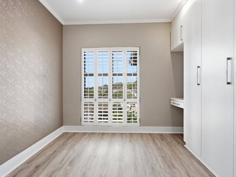 A bright, empty bedroom featuring modern laminate flooring, built-in cupboards with an integrated desk, and white plantation shutters.